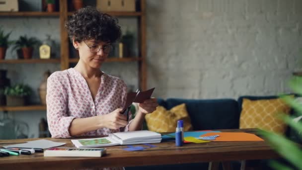 Giovane donna creativa sta tagliando pezzo di carta con le forbici creando collage poi mettendolo nel quaderno e controllando il design. Creatività, spazio di lavoro e concetto di arte . — Video Stock