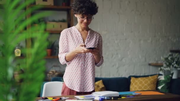 巻き髪の女性を笑顔はフラット デスクの写真を撮って置くモダンなアパートメントで立っているスマート フォンのカメラを使用しています。近代的な技術とデザインの概念. — ストック動画
