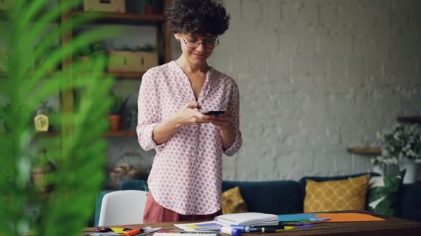 Glimlachend curly-haired dame is fotograferen van Bureau maken plat leggen met behulp van smartphone camera staande in modern appartement. Moderne technologie en design concept. — Stockvideo