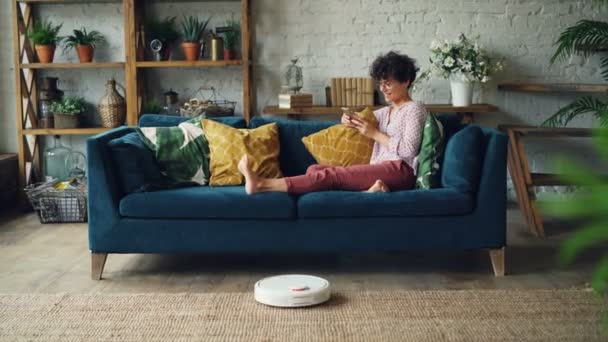 Glad tjej är att använda smartphone och leende sitter på soffan medan robotdammsugare städa golvet i modern lägenhet. Teknik och hushållsarbete koncept. — Stockvideo