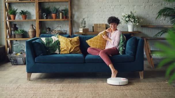 Joyeuse jeune femme joue avec l'aspirateur robotique puis en utilisant smartphone assis sur le canapé à la maison bénéficiant de la technologie moderne. Concept de personnes et d'innovations . — Video