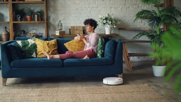 Estudante do sexo feminino está usando smartphone relaxante no sofá, enquanto hoover robótico está limpando chão em casa. Eletrodomésticos e conceito de tecnologia . — Vídeo de Stock