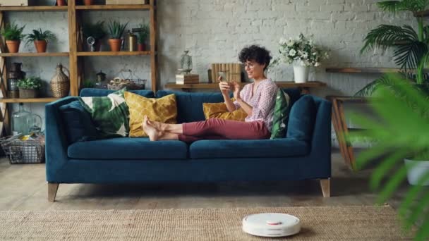 快乐的女孩手持智能手机触摸屏, 微笑着躺在沙发上, 而机器人吸尘器正在做家务清洁公寓地板. — 图库视频影像