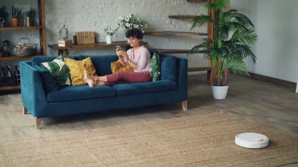 Sorrindo jovem dona da casa está sentado no sofá na sala de estar usando smartphone, enquanto hoover robótico está limpando o chão fazendo trabalho doméstico. Invenções e conceito de vida cotidiana . — Vídeo de Stock