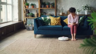 Çekici kadın kanepede oturan o zaman smartphone kullanarak ve aygıt evi temizlerken dinlenme robot elektrik süpürgesi üzerinde dönüyor. Teknoloji ve daireler kavramı.