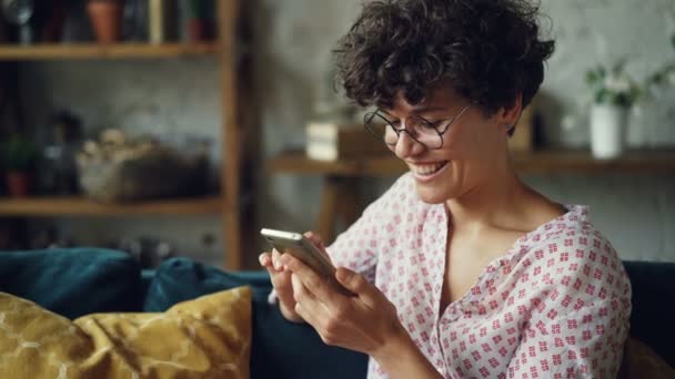 Happy girl is using smartphone looking at screen and laughing texting friends and checking social media accounts in internet sitting on sofa at home. — Stock Video