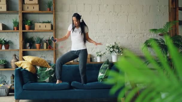 Carino ragazza asiatica sta ballando sul divano ascoltando musica in smartphone attraverso le cuffie e la firma godendo canzone all'interno in appartamento moderno. Gioventù e divertimento concetto . — Video Stock