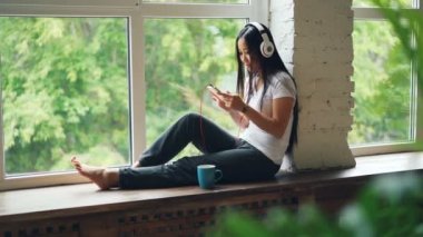 Şirin Asyalı kız esmer kulaklık müzik dinlemek ve smartphone dinlenme kullanarak pencere pervazına üzerinde oturuyor. Modern iç mekanlar, teknoloji ve insan kavramı.