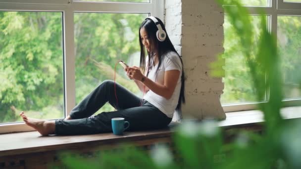 Cheerful Asian woman is listening to favourite song in headphones and using smartphone smiling and laughing sitting on window sill. Internet and youth culture concept. — Stock Video