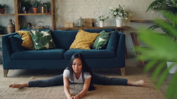 Flexible chica asiática está haciendo yoga en casa sentado en el suelo y estirando las piernas y el cuerpo haciendo ejercicio solo. Hermosa habitación con muebles y plantas está en el fondo . — Vídeo de stock