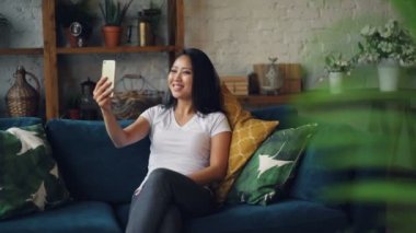 Modern Asya kız konuşuyor ve evde kanepede oturan ve arkadaşları ile iletişim zevk el hareketi sonra el sallayarak online video arama yapma.