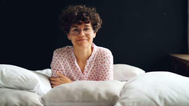 Αργή κίνηση πορτρέτο f ελκυστική σγουρά επικεφαλής γυναίκα γυαλιά και casual ένδυσης βλέπουν φωτογραφική μηχανή και χαμογελαστός ξαπλωμένη στο κρεβάτι μεταξύ μαλακά λευκά μαξιλάρια. — Αρχείο Βίντεο