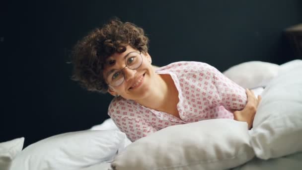 Portrait de fille joyeuse avec des cheveux bouclés courts portant des lunettes roulant dans le lit, souriant et regardant la caméra exprimant le bonheur. Chambre à coucher, émotions positives et concept jeunesse . — Video