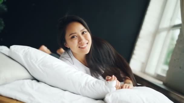Portrait of happy Asian lady in white T-shirt rolling in bed, smiling and looking at camera expressing positive emotions. Bedroom, joy and youth concept. — Stock Video