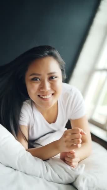 Slow motion of beautiful Asian brunette rolling in bed and looking at camera with glad smile having fun and enjoying herself. Apartment and millennials concept. — Stock Video