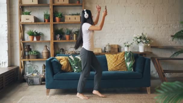 Movimento lento de mulher asiática bonita dançando em casa sozinha ouvindo música no smartphone através de fones de ouvido e cantando se divertindo. Conceito de tecnologia e apartamento . — Vídeo de Stock