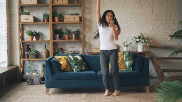 Slow motion van schattige Aziatische Dame dansen en springen op verdieping van moderne appartement dragen koptelefoon, houden van de smartphone en muziek beluisteren. Plezier en jeugd concept. — Stockvideo