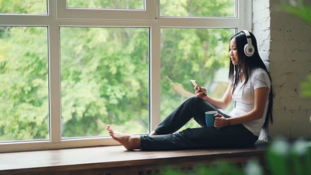 Relajada señora asiática está sentado en el alféizar de la ventana, escuchando música a través de auriculares inalámbricos y el uso de teléfono inteligente con sonrisa. Concepto de personas y ocio . — Vídeo de stock