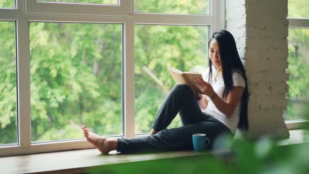 Attraktive asiatische Studentin liest Buch und sitzt lächelnd auf Fenstersims in einer modernen Wohnung. Hobby, Jugendkultur und Innenraumkonzept. — Stockvideo