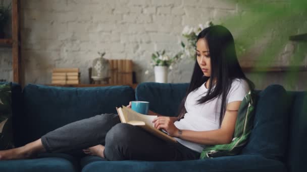 Bella donna asiatica sta leggendo libro godendo letteratura moderna seduta sul divano a casa in possesso di una tazza di tè e relax. Istruzione, millennials e concetto di casa . — Video Stock