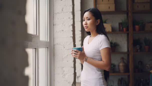 Smiling Asian woman is drinking tea standing near the window and looking outside enjoying beautiful view and leisure time at home. Apartments, drinks and people concept. — Stock Video