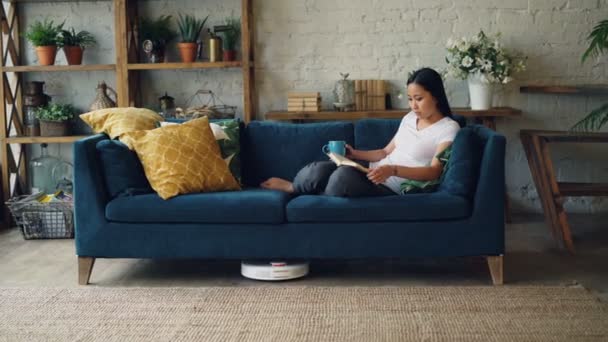 Hermosa chica asiática está leyendo libro descansando en el sofá en la sala de estar, mientras que la aspiradora robótica es piso de limpieza en seco y alfombra. Concepto de tecnología y personas . — Vídeo de stock