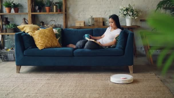 Расслабленная азиатка читает книгу, переворачивая страницы, сидя дома на диване, когда роботизированный пылесос чистит пол, выполняя домашнюю работу. Концепция отдыха и технологий . — стоковое видео