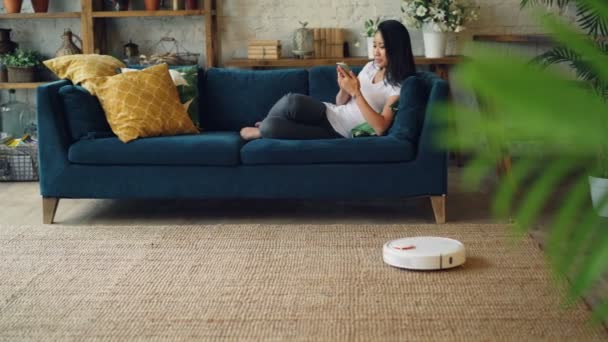 Estudante asiático está usando smartphone descansando no sofá quando hoover robótico é hoovering chão e carpete em casa. Eletrodomésticos e conceito de tecnologia . — Vídeo de Stock