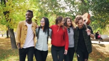 Slow motion of group of young men and women hipsters walking in park together with guitar laughing and talking. Youth lifestyle and autumn nature concept.