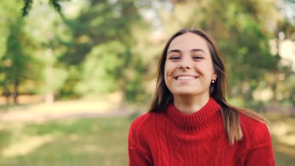 Retrato en cámara lenta de chica alegre en suéter caliente de pie en el parque con sonrisa alegre, riendo y mirando a la cámara. Naturaleza y concepto de los millennials . — Vídeo de stock