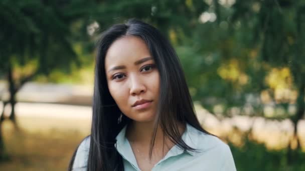 Retrato en cámara lenta de una chica asiática guapa con el pelo largo usando una camisa azul claro de pie en el parque, sonriendo y mirando a la cámara. Naturaleza y concepto de juventud . — Vídeo de stock