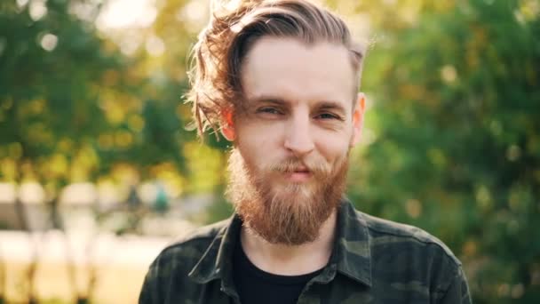 Slow motion portrait of happy bearded guy hipster in military style clothing standing in the park smiling and looking at camera. People and emotions concept. — Stock Video