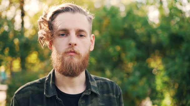 Slow motion portret van ernstige bebaarde jongeman in militaire stijl shirt permanent in het park op herfstdag en camera te kijken. Mens en natuur concept. — Stockvideo