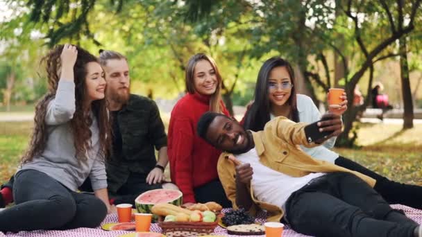 Movimento lento de homem afro-americano feliz tomando selfie com amigos no piquenique usando smartphone, as pessoas estão posando com bebidas e mostrando gestos de mão . — Vídeo de Stock