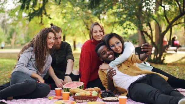 Zeitlupe fröhlicher junger Hipster beim Picknick mit dem Smartphone, Männer und Frauen posieren, umarmen und zeigen Handgesten. — Stockvideo