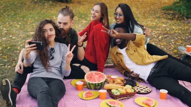 Mouvement lent des filles et des gars insouciants groupe multiethnique prenant selfie avec smartphone assis sur plaid dans le parc avec de la nourriture et des boissons s'amuser ensemble . — Video
