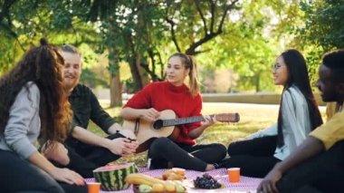 Ağır çekim neşeli insanlar şarkı ve güzel genç kadın parkta piknik sırasında gitar çalarken gülüyor. Müzik, eğlence ve eğlence kavramı.