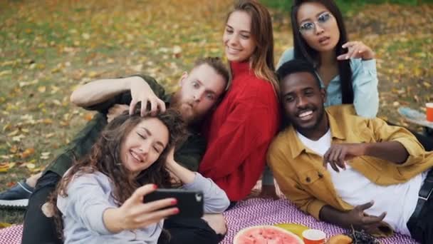 Slow motion van vrolijke mannen en vrouwen nemen selfie met smartphone en plezier liggend op de deken in het park in het najaar. Picknick, aard en gadgets concept. — Stockvideo