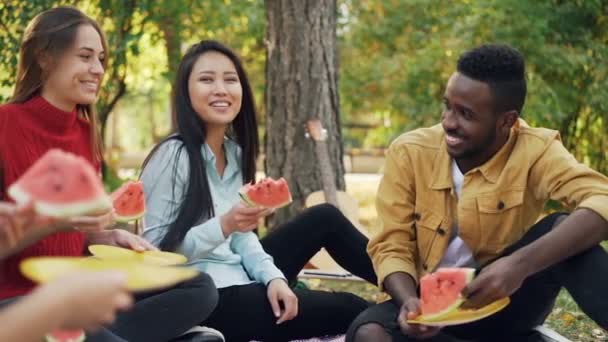 Rallentatore di giovani allegri che mangiano anguria, parlano e ridono su picnic in parco durante giorno di autunno. Amicizia, cibo e comunicazione . — Video Stock