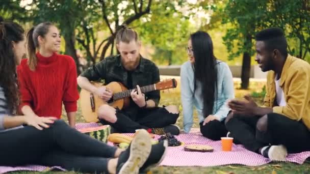 Hipster alegre está cantando e tocando guitarra sentado em cobertor no parque com os amigos e se divertindo, as pessoas estão batendo palmas. Conceito de comida, música e diversão . — Vídeo de Stock