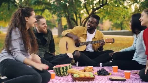 Vrolijke Afro-Amerikaanse man speelt de gitaar voor zijn vrienden zittend op een deken op picknick en het genieten van de warme herfst weekend. Vrije tijd en muziek concept. — Stockvideo