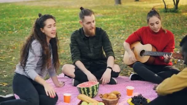 Alunos alegres estão desfrutando de música sentados em cobertor no parque e tocando as músicas de canto de guitarra no piquenique. Tempo agradável com amigos, natureza e conceito divertido . — Vídeo de Stock