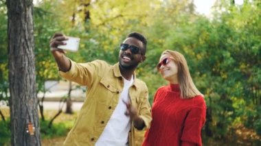 Güzel çift Afro-Amerikan erkek ve beyaz kadın akıllı telefon tutarak ve poz parkta selfie alıyor v-sign ve dil gösteriliyor. Fotoğraf ve insanlar kavramı.
