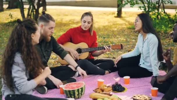 Menina bonita está tocando guitarra enquanto seus amigos estão cantando e ouvindo música descansando em xadrez no parque. Frutas alimentares e pastelaria são visíveis no cobertor . — Vídeo de Stock