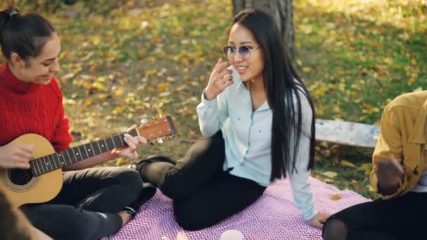 Amici gioiosi multi-etnico gruppo stanno cantando canzoni seduti sulla coperta nel parco, mentre la bella donna sta suonando la chitarra. Natura, millennials e concetto musicale . — Video Stock