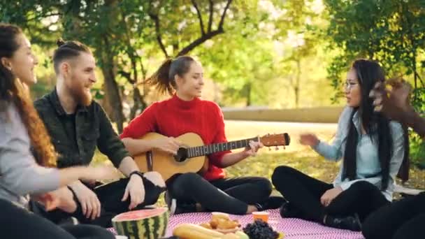 Les jeunes gens ludiques chantent et bougent les mains lorsque la belle fille joue de la guitare pendant le pique-nique dans le parc le jour ensoleillé d'automne. Concept amusant et musical . — Video