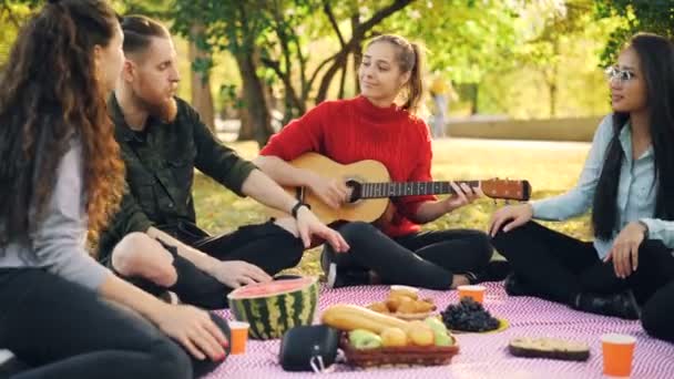 Los estudiantes relajantes están haciendo un picnic en el parque tocando canciones de guitarra y disfrutando del tiempo libre al aire libre con amigos. La comida en la manta y la naturaleza es visible . — Vídeo de stock