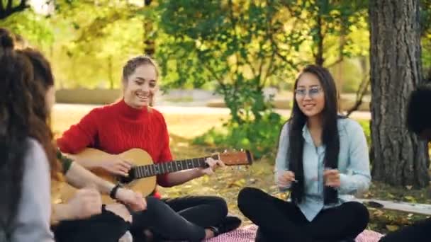 Guitarrista feminina está tocando a guitarra para amigos alegres sentados em cobertor no gramado no parque da cidade, enquanto caras e meninas estão ouvindo, rindo e se divertindo . — Vídeo de Stock