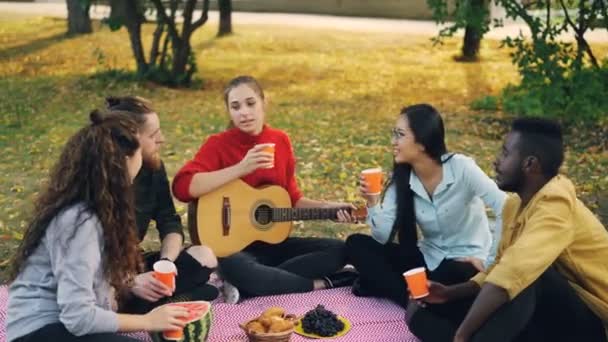 Jóvenes alegres grupo multiétnico son tintinear vasos con bebidas y luego beber sentado en la manta en la hierba durante el picnic de otoño con la guitarra y la comida . — Vídeo de stock