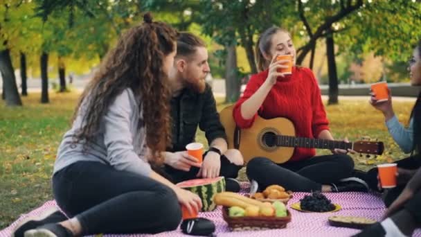 Молоді чоловіки і жінки готують тости і клінкерні окуляри на пікніку в парку з гітарою в теплий осінній день. Концепція дружби, напоїв та дозвілля . — стокове відео